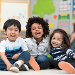 three toddlers smile in a brightly colored classroom